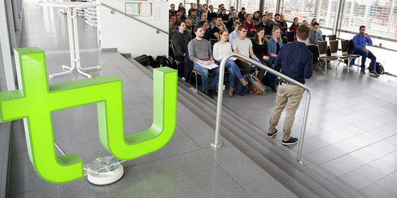 A large hall with a window front in which a group of about 50 young people are sitting on chairs listening to a lecture. In the foreground is a large TU logo glowing green.