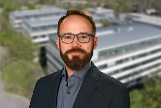 Porträtfoto von Prof. Lauven: Ein Mann mit Brille und Bart lächelt in die Kamera. Im Hintergrund ist ein verschwommenes Foto des Campus der TU Dortmund.