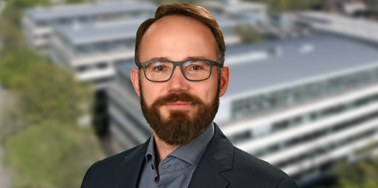 Porträtfoto von Prof. Lauven: Ein Mann mit Brille und Bart lächelt in die Kamera. Im Hintergrund ist ein verschwommenes Foto des Campus der TU Dortmund.
