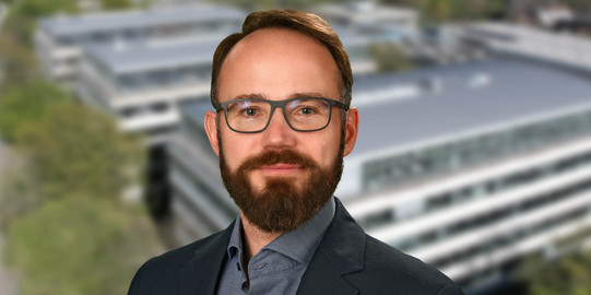 Portrait photo of Prof. Lauven: A man with glasses and a beard smiles into the camera. In the background is a blurred photo of the TU Dortmund campus.