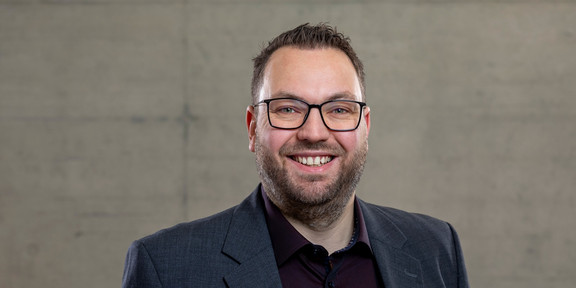 Foto: Ein Mann mit kurzem dunklem Haar steht vor einer grauen Wand und lächelt in die Kamera.