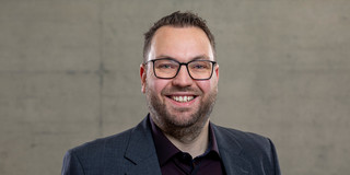 Foto: Ein Mann mit kurzem dunklem Haar steht vor einer grauen Wand und lächelt in die Kamera.