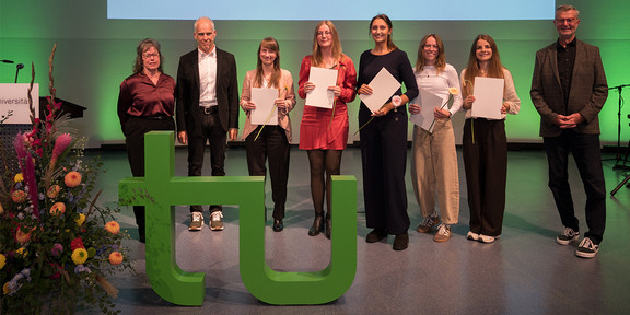 Foto: Acht Personen stehen auf einer grün beleuchteten Bühne vor einer Leinwand mit Titeltext, fünf von ihnen halten Papiere und Blumen, im Vordergrund steht ein grünes „tu"-Logo.