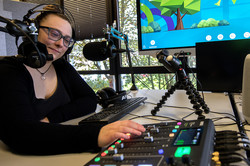 Eine Frau mit Kopfhörern sitzt in einem Tonstudio. Vor ihr stehen ein Mischpult, ein Mikrophon sowie Tastatur und Kopfhörer. An der Wand ist ein Monitor zu erkennen.
