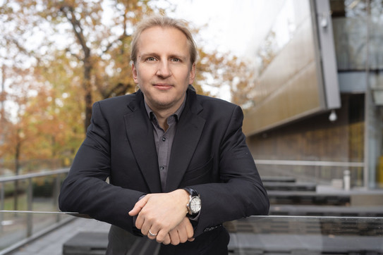Portrait photo: Prof. Michael Kamp wears a dark suit and leans on a parapet.