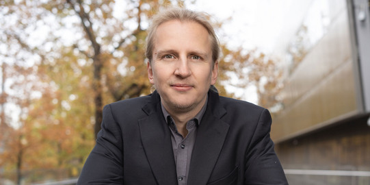 Portrait photo: Prof. Michael Kamp wears a dark suit and leans on a parapet.
