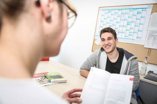 Ein Student sitzt gegenüber von einer Frau welche ein Buch liest