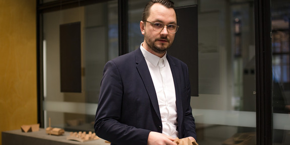 Felix Lowin Ein Mann im Anzug steht vor einer Glasfront und hält ein Holzbaustein in dem Händen