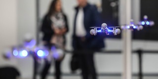 In the foreground four drones are flying, in the background two people can be seen