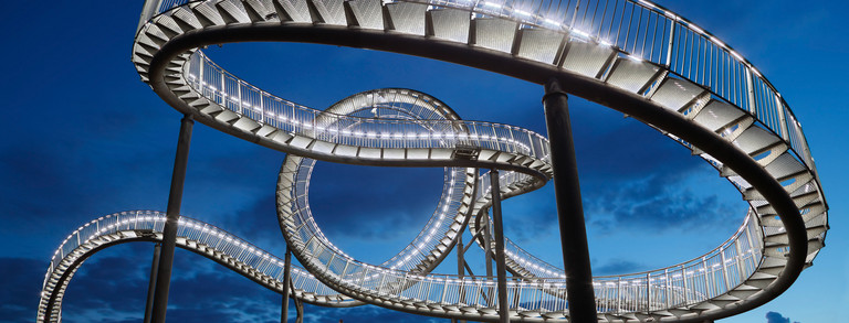 Illuminated staircase sculpture “Tiger and Turtle,” designed to resemble a roller coaster, against the evening sky