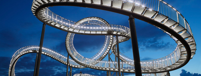 Illuminated staircase sculpture “Tiger and Turtle,” designed to resemble a roller coaster, against the evening sky