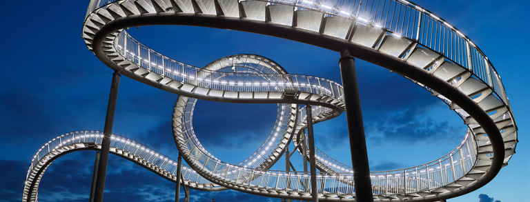 Beleuchtete Treppenskulptur "Tiger and Turtle", ähnlich wie eine Achterbahn gestaltet, vor abendlichem Himmel