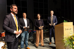 Drei Männer und eine Frau in Anzügen stehen im Halbkreis auf einer Bühne. Einer der Männer spricht.