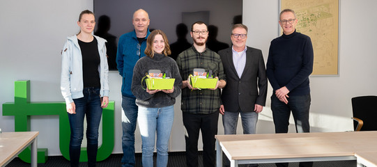 Foto: Eine Gruppe von Personen steht in einem Raum, zwei von ihnen halten Geschenkkörbe in den Händen.