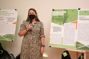 A woman stands next to a poster and speaks into the microphone