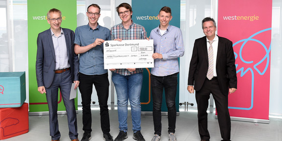 Das Siegerteam des Westenergie Manager Cups Gruppenfoto von fünf Männern im Business-Outfit vor drei Werbebannern. Links steht Prof. Andreas Hoffjan, in der Mitte stehen drei Studenten und halten einen großen Sparkassen-Check, rechts steht Achim Schröder.
