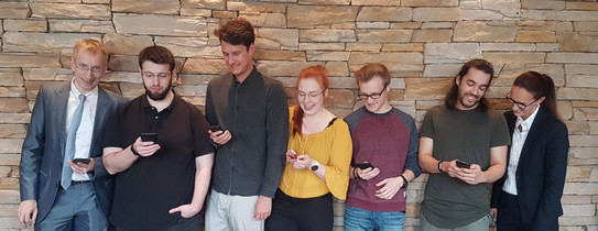 Sieben Personen stehen vor einer Wand und haben ihre Handys in der Hand