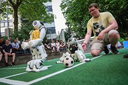 Vorne auf einem grünen Teppich-Fußballfeld sind zwei ca. 50 Centimeter große Roboter. Der linke trägt ein gelbes Trikot mit schwarzer Schrift und steht links von einem kleinen Fußball. Der rechte trägt ein schwarzen Trikot mit gelber Schrift und steht gerade vom Sitzen auf. Der Ball liegt vor ihm. Hinter ihm hockt ein Student mit Brille und gelbem T-Shirt, der seine Hand hinter dem Roboter zur Unterstützung bereit hält. Im Hintergrund sitzt ein Publikum auf Außenbänken und schaut zu.