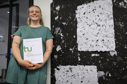 Foto: Eine junge Frau mit blonden Haaren steht vor einem Kunstwerk und hält eine Urkunde in den Händen.