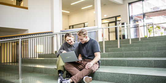 Studieninteressierte Ein Foto mit zwei Studieninteressierten, die lächelnd auf einer Treppe in der TU Dortmund sitzen und sich gemeinsam Informationen auf einem Laptop anschauen.