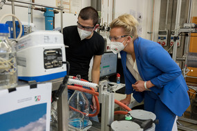 Ina Brandes und ein Mann stehen mit Schutzbrille und Schutzmaske vor einer Apparatur in einem Labor.