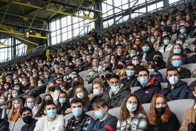 Auf einer überdachten Tribüne in einem Fußballstadion sitzen Erstsemesterstudierende, die eine Mund-Nasen-Bedeckung tragen.