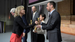 Eine Frau überreicht einem Mann einen schwarz-goldene Trophäe auf einer Bühne.