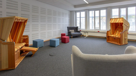 Raum mit zwei Strandkörben aus Holz und Stoff, mehreren Sitzwürfeln und Sofas vor großen Fenstern