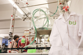 Ein weißer Laborkittel mit der grünen Aufschrift "bci" hängt vor einer Apparatur aus Kolben und Schläuchen.