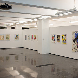 Das Bild zeigt einen Ausstellungsraum mit verschiedenen internationalen Künstlerplakaten an einer weißen Wand.