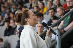 Eine Frau hält ein Mikrofon und schaut nach oben. Im Hintergrund eine volle Tribüne.