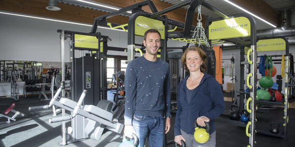 A man and a woman stand in front of a fitness machine and smile at the camera.