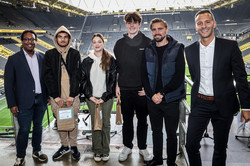 Gruppenbild von sechs Personen. Drei Studierende werden links und rechts von den Veranstaltenden flankiert. Alle lächeln in die Kamera. Im Hintergrund das Stadion.