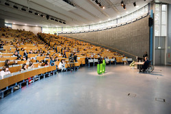 Ein großer Hörsaal von der Bühne aus. Rechts im Bild sitzen zwei Männer an einem Tisch. Zwischen ihnen und den Sitzreihen steht ein großes TU-Logo in grün. In den Sitzreihen sitzen Menschen verschiedener Generationen und hören zu.