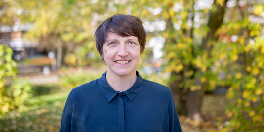 Porträtfoto: Ein Porträtfoto von Prof. Hannah Zagel vor Bäumen auf dem Campus Nord. Hannah Zagel hat kurze dunkle Haare und trägt eine dunkelblaue Bluse. Die Bäume im Hintergrund tragen gelbes Laub.