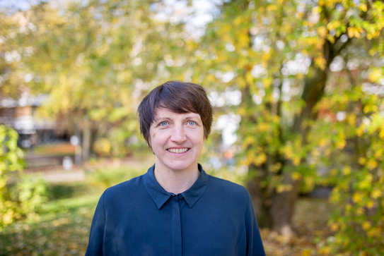 Porträtfoto: Ein Porträtfoto von Prof. Hannah Zagel vor Bäumen auf dem Campus Nord. Hannah Zagel hat kurze dunkle Haare und trägt eine dunkelblaue Bluse. Die Bäume im Hintergrund tragen gelbes Laub.