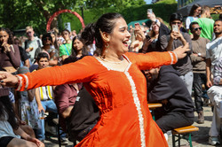Foto: Eine Frau mit dunklen Haaren und orangem Kleid tanzt mit ausgebreiteten Armen auf dem Sommerfest. Im Hintergrund sitzen und stehen Menschen im Publikum.