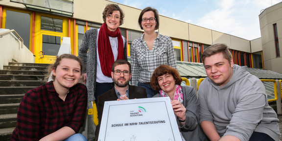 Four people squat on the floor and hold a plaque. Behind them stand two women.