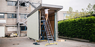 Eine Person arbeitet auf einer Leiter an einem unvollständigen Tiny-House. Das Gebäude hat grüne Wände und ein flaches Dach. Es steht auf Betonboden. 