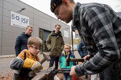 Im Vordergrund des Bildes ist ein Kind mit Schutzbrille zu sehen, welches mit einem Hammer auf einen Metallring schlägt. Der Metallring wird von einem Mann mit Zange gehalten. Im Hintergrund sind weitere Personen zu sehen, die in die Richtung des Jungen gucken sowie ein grauses Gebäude, an dem zwei Plakate hängen.