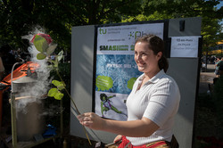 Eine junge Frau steht an einem Tisch mit Plexiglas-Scheibe und hält in der einen Hand eine tiefgefrorene Rose und einen Hammer in der anderen.