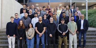 Auf dem Foto sieht man die neuen Auszubildenden für ein Gruppenfoto in die Kamera lächeln. Sie stehen vor einer Treppe auf dem Campus Süd.