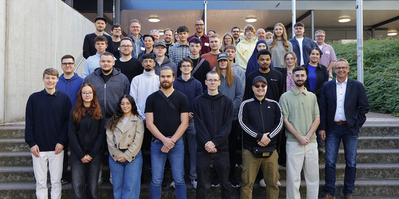 In the photo, the new trainees are smiling at the camera for a group photo. They are standing in front of a staircase on the South Campus.