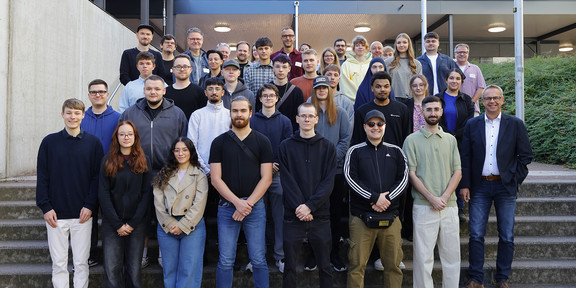 Auf dem Foto sieht man die neuen Auszubildenden für ein Gruppenfoto in die Kamera lächeln. Sie stehen vor einer Treppe auf dem Campus Süd.