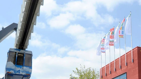 H-Bahn neben rotem Gebäude mit mehreren wehenden internationalen Flaggen im Wind, umgeben von Bäumen