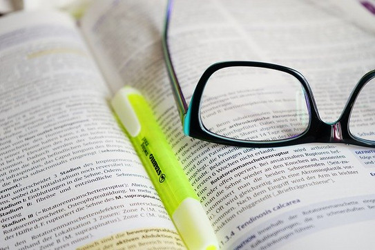 Genaues Lesen In Nahaufnahme ist ein aufgeschlagenes Buch zu sehen, auf diesem liegen Stift und Brille.