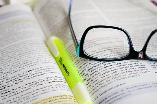 Genaues Lesen In Nahaufnahme ist ein aufgeschlagenes Buch zu sehen, auf diesem liegen Stift und Brille.