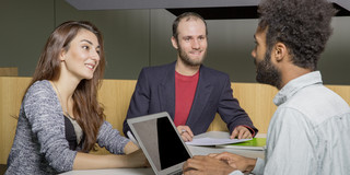Ein junger Mann sitzt an einem Laptop, während ein weiterer junger Mann und eine junge Frau ihm gegenübersitzen.