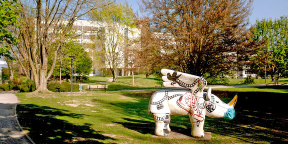 A large white sculpture of a winged rhinoceros, with a mouth guard on, stands in a meadow. In the background you can see large white buildings.