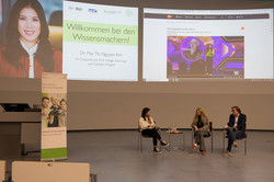 Three people are sitting on chairs in conversation. A presentation is playing in the background.